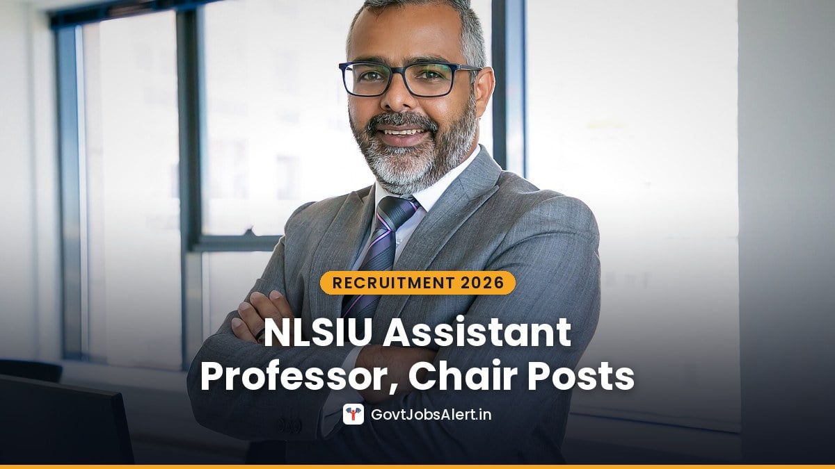Smiling professor in a grey suit with arms crossed in an office, NLSIU Recruitment 2026 for Assistant Professor and Chair posts